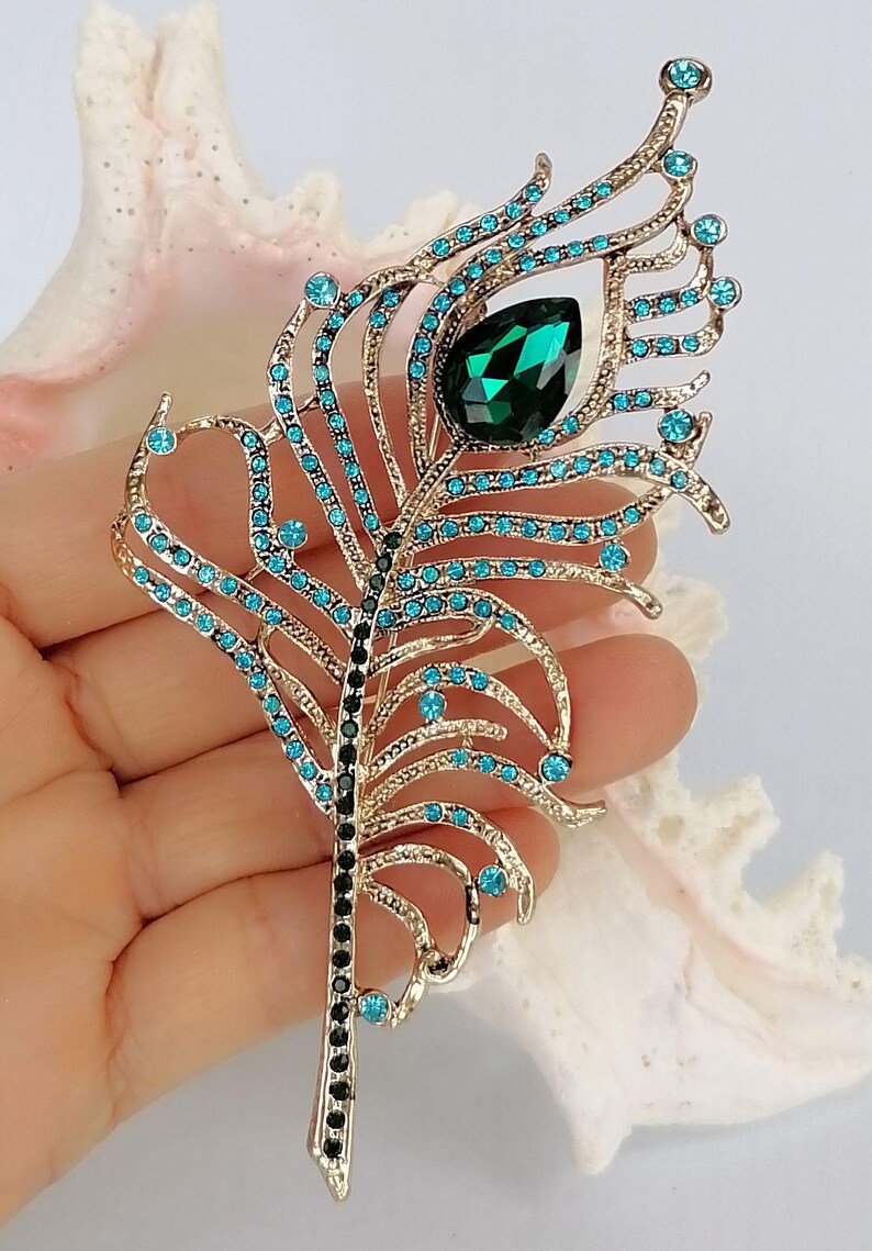 May include: Gold-coloured metal peacock feather brooch with teal and black accents. The brooch features teal-coloured rhinestones and a large emerald-green teardrop-shaped stone. A decorative accessory.