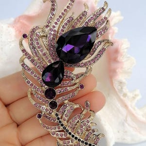May include: A gold-toned metal brooch shaped like a feather, embellished with numerous small light purple rhinestones and two large, prominent, dark purple teardrop-shaped crystals positioned along the central spine of the feather design.