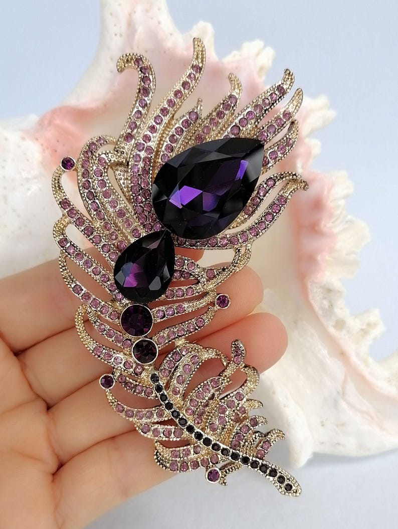 May include: A gold-toned metal brooch shaped like a feather, embellished with numerous small light purple rhinestones and two large, prominent, dark purple teardrop-shaped crystals positioned along the central spine of the feather design.