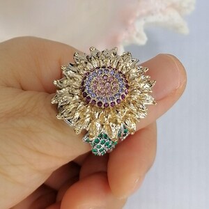 K&ouml;nnte beinhalten: Ein goldfarbener Ring in Form einer Sonnenblume mit strukturierten Bl&uuml;tenbl&auml;ttern. Die Mitte besteht aus konzentrischen Kreisen aus kleinen rosa, hellvioletten und dunkelvioletten Steinen. Ein Blattakzent ist mit gr&uuml;nen Steinen besetzt.
