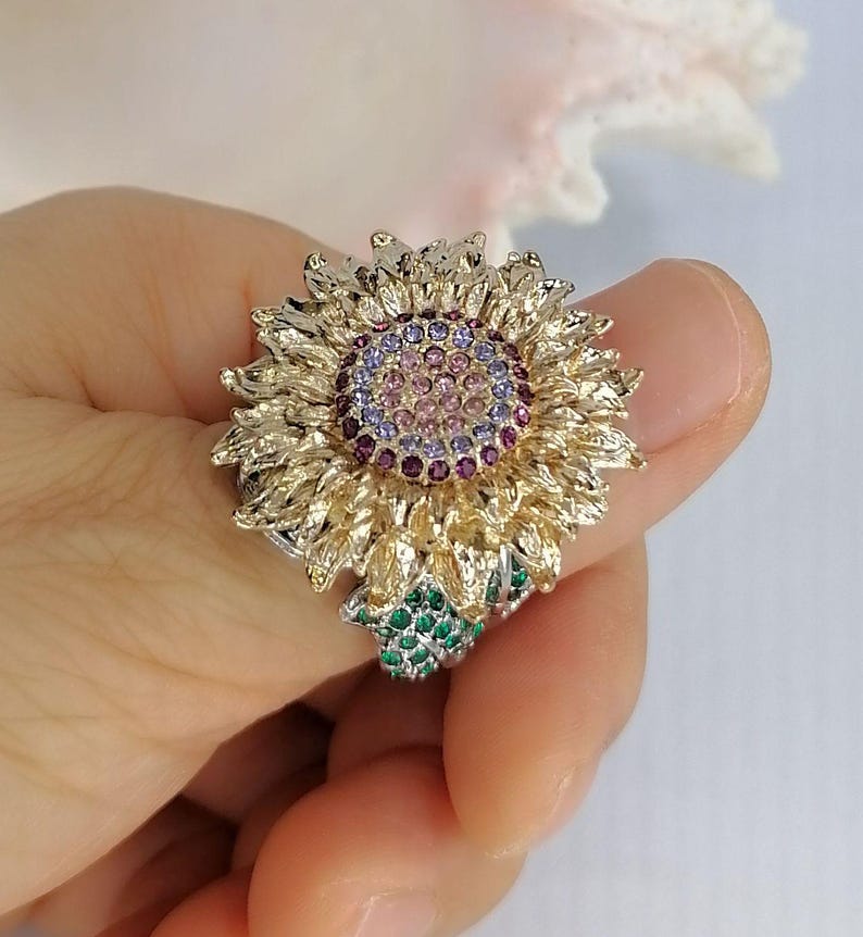 K&ouml;nnte beinhalten: Ein goldfarbener Ring in Form einer Sonnenblume mit strukturierten Bl&uuml;tenbl&auml;ttern. Die Mitte besteht aus konzentrischen Kreisen aus kleinen rosa, hellvioletten und dunkelvioletten Steinen. Ein Blattakzent ist mit gr&uuml;nen Steinen besetzt.