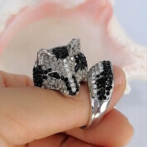 Pu&ograve; includere: Un anello color argento a forma di testa di procione con un design a coda avvolgente. La superficie &egrave; ricoperta da piccoli cristalli neri e trasparenti scintillanti che formano i tratti del viso e il motivo a strisce della coda.