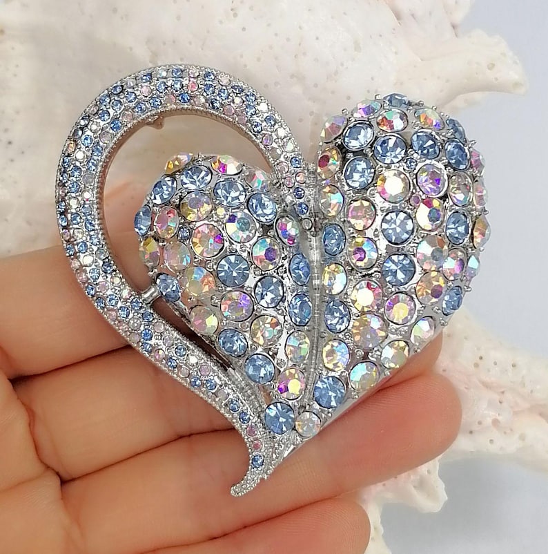 May include: A heart-shaped brooch with a silver-tone metal frame. The brooch is adorned with light blue, iridescent, and pink rhinestones. The brooch is held in a hand.