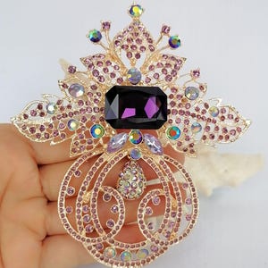 May include: A gold-tone metal brooch featuring a large rectangular dark purple central stone, surrounded by intricate filigree patterns adorned with small light purple crystals and iridescent round stones, including a dangling teardrop-shaped charm.