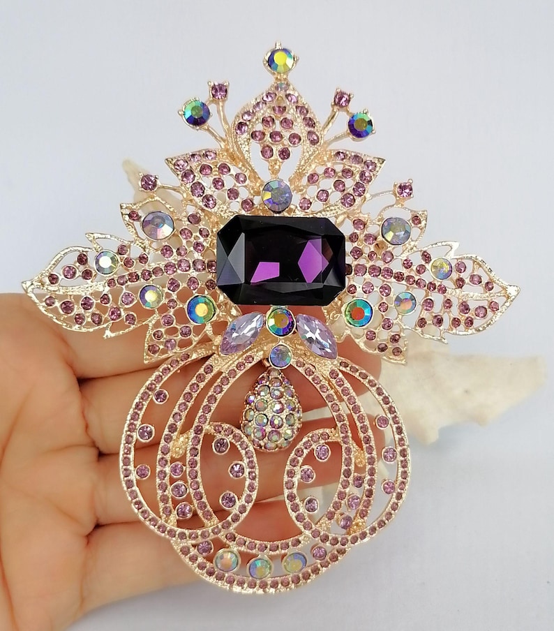 May include: A gold-tone metal brooch featuring a large rectangular dark purple central stone, surrounded by intricate filigree patterns adorned with small light purple crystals and iridescent round stones, including a dangling teardrop-shaped charm.