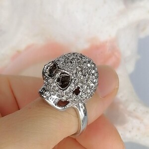 Op de afbeelding: Een zilverkleurige ring met een driedimensionaal schedelontwerp. Het gehele oppervlak van de schedel is ingelegd met talloze kleine, heldere, fonkelende steentjes. De ring heeft een gladde, gepolijste zilveren band en donkere, diepe oogkassen.