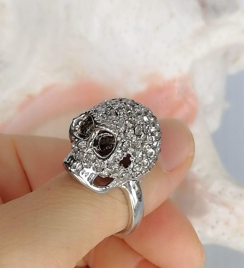 Op de afbeelding: Een zilverkleurige ring met een driedimensionaal schedelontwerp. Het gehele oppervlak van de schedel is ingelegd met talloze kleine, heldere, fonkelende steentjes. De ring heeft een gladde, gepolijste zilveren band en donkere, diepe oogkassen.