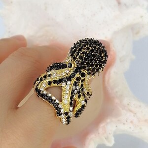 Peut inclure: Une bague dor&eacute;e en forme de pieuvre, dont la t&ecirc;te et les tentacules sont incrust&eacute;s de nombreuses petites pierres &eacute;tincelantes noires et blanches transparentes. La base m&eacute;tallique est visible entre les pierres, cr&eacute;ant une surface textur&eacute;e.