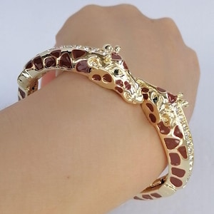 May include: A gold-tone hinged bangle bracelet featuring two giraffe heads meeting in the center. The bracelet is decorated with brown enamel giraffe spots and small clear crystals along the sides of the band, creating a detailed animal motif.
