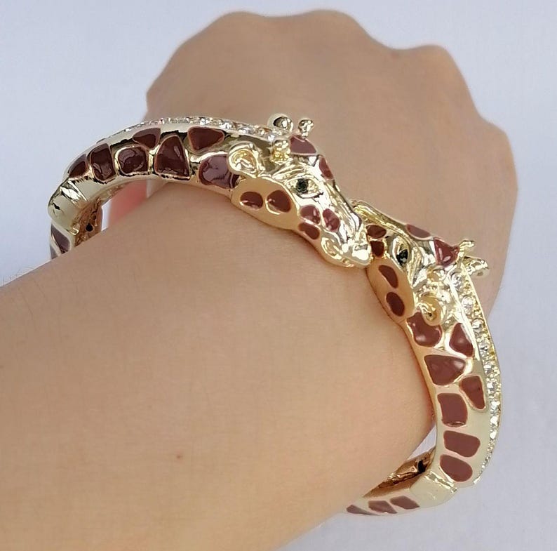 May include: A gold-tone hinged bangle bracelet featuring two giraffe heads meeting in the center. The bracelet is decorated with brown enamel giraffe spots and small clear crystals along the sides of the band, creating a detailed animal motif.