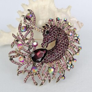 May include: A gold-toned unicorn brooch with a detailed design. The unicorn's body is covered in small, deep purple crystals, with a large, teardrop-shaped, dark red gem at the base. The brooch is accented with pink and iridescent crystals.