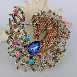 May include: A gold-toned seahorse brooch encrusted with colorful rhinestones. The seahorse features a brown body, a large blue teardrop-shaped gem, and accents of pink, green, and other colors. The brooch is set against a white background.