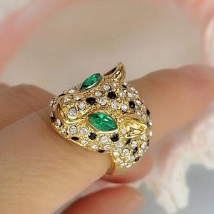 May include: A gold-toned ring shaped like a leopard head, featuring two marquise-cut green stones for eyes. The surface is covered in small, clear white crystals and scattered black crystals, creating a spotted pattern across the metal head.