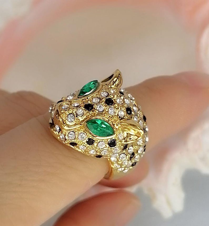 May include: A gold-toned ring shaped like a leopard head, featuring two marquise-cut green stones for eyes. The surface is covered in small, clear white crystals and scattered black crystals, creating a spotted pattern across the metal head.