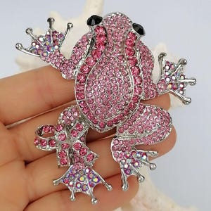 K&ouml;nnte beinhalten: Ein pinkfarbener Strassfrosch-Broche mit silberfarbenem Metallsockel. Der Froschk&ouml;rper ist mit kleinen, pinkfarbenen Strasssteinen bedeckt, mit schwarzen Strassaugen. Die Brosche hat ein detailliertes Design.