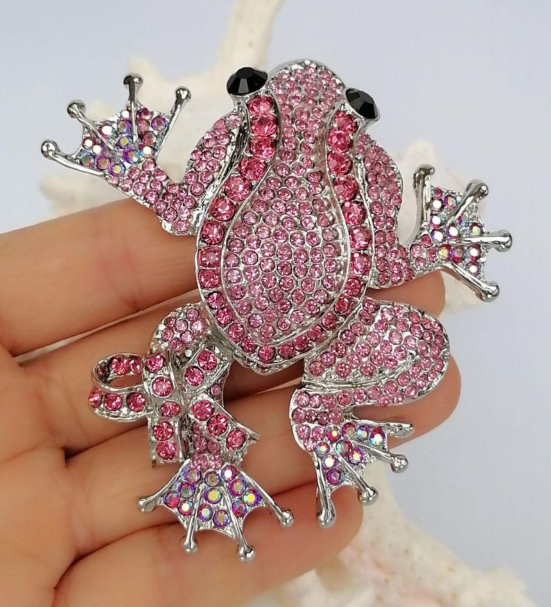 K&ouml;nnte beinhalten: Ein pinkfarbener Strassfrosch-Broche mit silberfarbenem Metallsockel. Der Froschk&ouml;rper ist mit kleinen, pinkfarbenen Strasssteinen bedeckt, mit schwarzen Strassaugen. Die Brosche hat ein detailliertes Design.