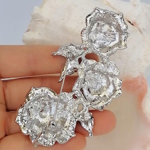 May include: A silver-toned metal brooch featuring three rose blossoms arranged in a vertical cluster. The back of the piece displays a textured metallic finish with a long, thin pin mechanism attached for fastening, showing the reverse side construction.