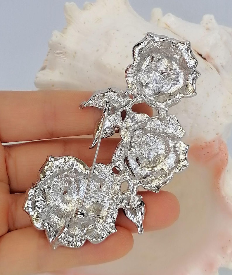 May include: A silver-toned metal brooch featuring three rose blossoms arranged in a vertical cluster. The back of the piece displays a textured metallic finish with a long, thin pin mechanism attached for fastening, showing the reverse side construction.