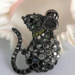 May include: A dark gray mouse-shaped brooch, encrusted with various sizes of gray and green rhinestones. The brooch has a curved tail and a small, round head. The background is a soft, blurred white and pink.