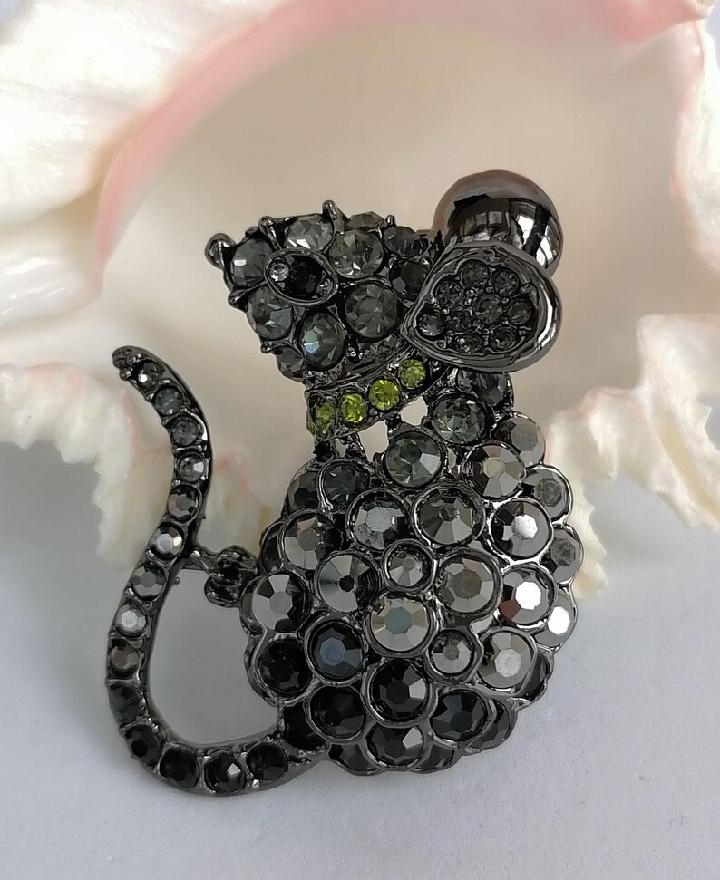 May include: A dark gray mouse-shaped brooch, encrusted with various sizes of gray and green rhinestones. The brooch has a curved tail and a small, round head. The background is a soft, blurred white and pink.