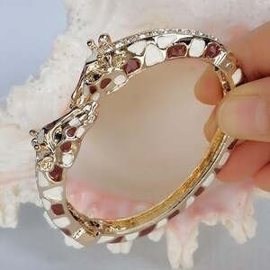May include: A hinged bangle bracelet featuring two giraffe heads meeting at the center. The bracelet is crafted from gold-tone metal with white and brown enamel patches covering the entire surface, mimicking a giraffe's natural coat pattern.