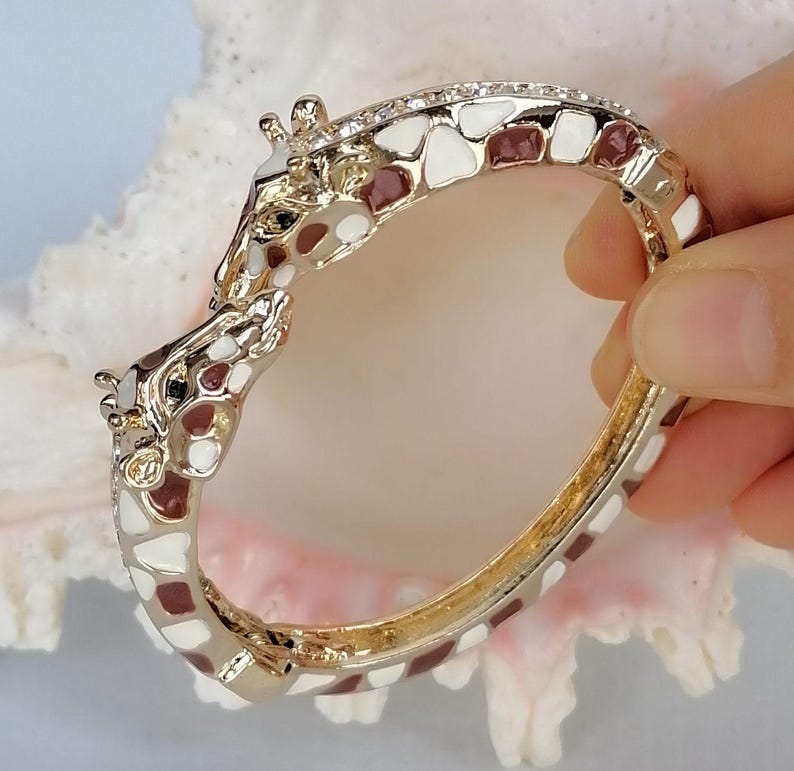 May include: A hinged bangle bracelet featuring two giraffe heads meeting at the center. The bracelet is crafted from gold-tone metal with white and brown enamel patches covering the entire surface, mimicking a giraffe's natural coat pattern.
