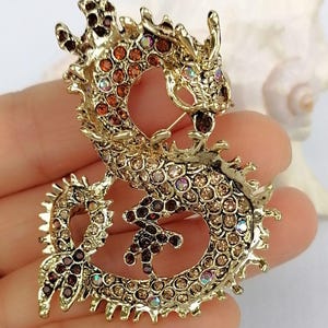 Puede incluir: Broche de drag&oacute;n dorado con brillantes pedrer&iacute;a en tonos marr&oacute;n, &aacute;mbar e iridiscentes. El drag&oacute;n est&aacute; intrincadamente detallado con un cierre de alfiler en la parte posterior. Un accesorio decorativo para chaquetas, bufandas o bolsos.