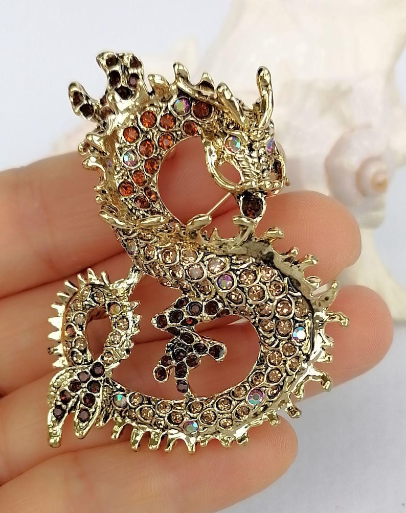 Puede incluir: Broche de drag&oacute;n dorado con brillantes pedrer&iacute;a en tonos marr&oacute;n, &aacute;mbar e iridiscentes. El drag&oacute;n est&aacute; intrincadamente detallado con un cierre de alfiler en la parte posterior. Un accesorio decorativo para chaquetas, bufandas o bolsos.