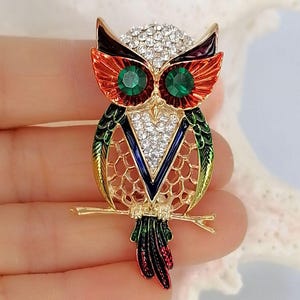 Op de afbeelding: Een uilbroche met een goudkleurig metalen frame en levendige emaille accenten in rood, oranje, groen en blauw. De uil is versierd met heldere strass-steentjes op de kop en borst, en twee grote, ronde, gefacetteerde groene stenen dienen als ogen.