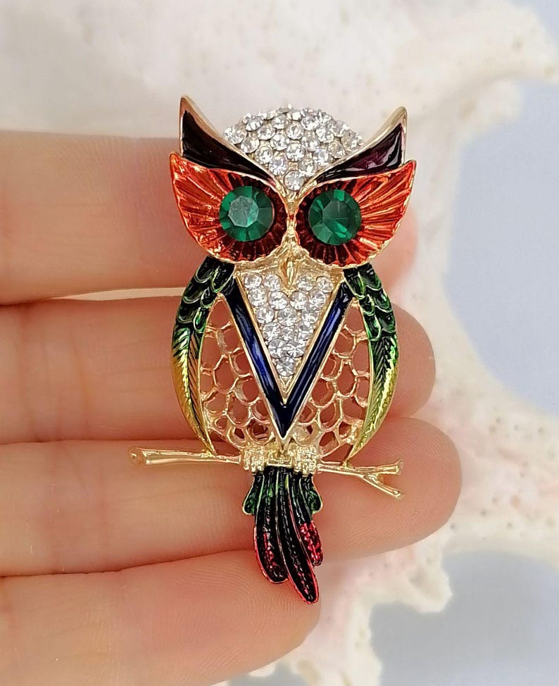 Op de afbeelding: Een uilbroche met een goudkleurig metalen frame en levendige emaille accenten in rood, oranje, groen en blauw. De uil is versierd met heldere strass-steentjes op de kop en borst, en twee grote, ronde, gefacetteerde groene stenen dienen als ogen.
