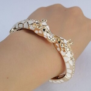 May include: A gold-toned hinged bangle bracelet featuring two giraffe heads meeting at the center. The giraffe bodies are decorated with white enamel patches and small clear crystals along the sides of the band, creating a detailed animal motif.