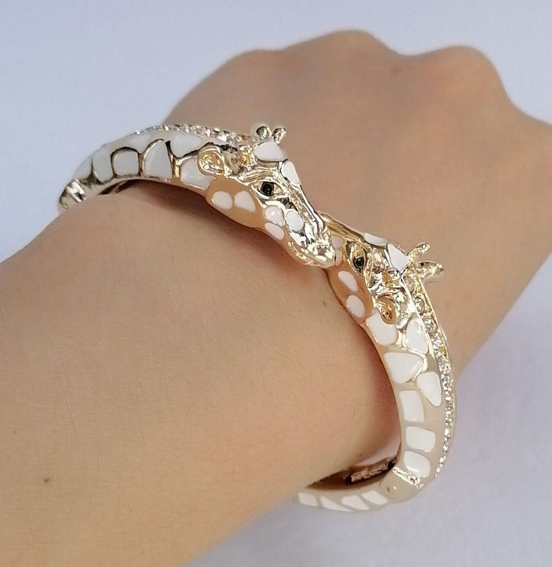 May include: A gold-toned hinged bangle bracelet featuring two giraffe heads meeting at the center. The giraffe bodies are decorated with white enamel patches and small clear crystals along the sides of the band, creating a detailed animal motif.