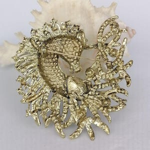 May include: A gold-toned seahorse brooch with intricate detailing. The seahorse is surrounded by ornate, textured embellishments. The brooch has a pin closure on the back. The background is a white seashell.
