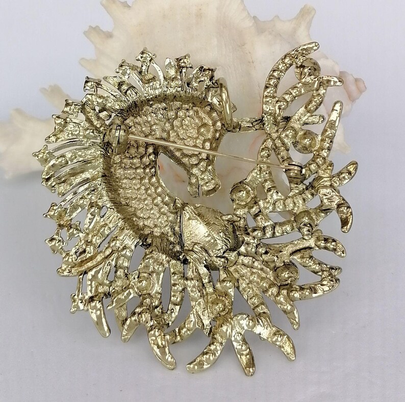 May include: A gold-toned seahorse brooch with intricate detailing. The seahorse is surrounded by ornate, textured embellishments. The brooch has a pin closure on the back. The background is a white seashell.