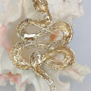 May include: A gold-coloured snake brooch with a textured finish. The snake is coiled, with its head raised and a pin on the back for fastening. The brooch is set against a white background.