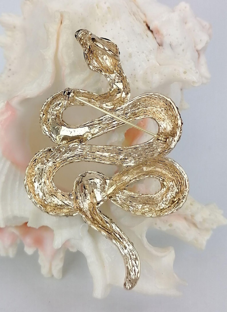 May include: A gold-coloured snake brooch with a textured finish. The snake is coiled, with its head raised and a pin on the back for fastening. The brooch is set against a white background.