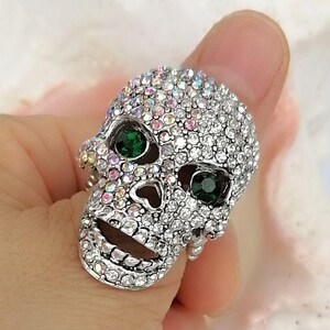 May include: A silver-toned ring shaped like a human skull, entirely encrusted with small, iridescent clear crystals. The skull features two prominent, round, dark green faceted stones set into the eye sockets, creating a striking contrast.