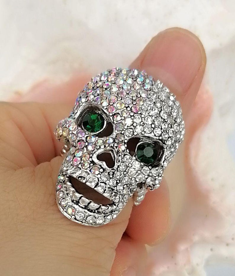 May include: A silver-toned ring shaped like a human skull, entirely encrusted with small, iridescent clear crystals. The skull features two prominent, round, dark green faceted stones set into the eye sockets, creating a striking contrast.