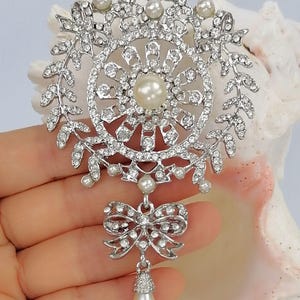 May include: A silver-toned brooch featuring a circular frame adorned with clear rhinestones and small white pearls. The design includes a central pearl, leaf-like side accents, and a dangling bow element with a single teardrop-shaped pearl pendant.