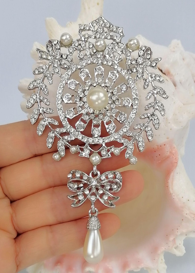 May include: A silver-toned brooch featuring a circular frame adorned with clear rhinestones and small white pearls. The design includes a central pearl, leaf-like side accents, and a dangling bow element with a single teardrop-shaped pearl pendant.
