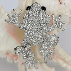 K&ouml;nnte beinhalten: Silberfarbene Froschbrosche, besetzt mit klaren Strasssteinen und schwarzen Edelsteinaugen. Der K&ouml;rper des Frosches ist mit kleinen, funkelnden Steinen bedeckt, mit detaillierten F&uuml;&szlig;en und ausgestreckten Armen. Die Brosche ist vor einem wei&szlig;en und rosa Muschelschalenhintergrund platziert.