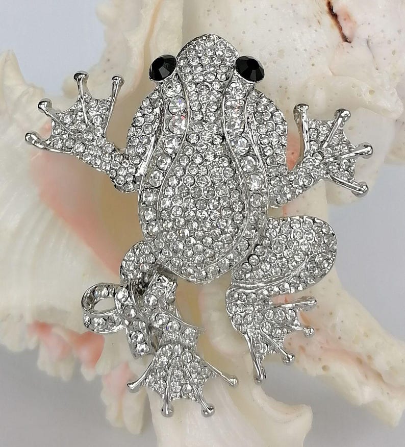 K&ouml;nnte beinhalten: Silberfarbene Froschbrosche, besetzt mit klaren Strasssteinen und schwarzen Edelsteinaugen. Der K&ouml;rper des Frosches ist mit kleinen, funkelnden Steinen bedeckt, mit detaillierten F&uuml;&szlig;en und ausgestreckten Armen. Die Brosche ist vor einem wei&szlig;en und rosa Muschelschalenhintergrund platziert.