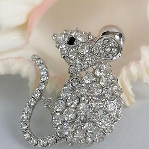 May include: A silver-toned mouse brooch encrusted with clear, sparkling rhinestones. The brooch features a detailed design of a mouse with a curved tail and a large, round head. The mouse's eye is a small black stone. The brooch is set against a white and pink background.