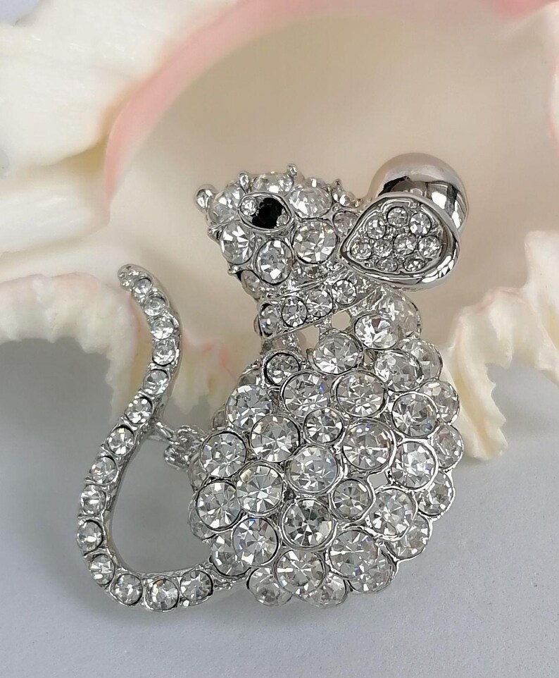 May include: A silver-toned mouse brooch encrusted with clear, sparkling rhinestones. The brooch features a detailed design of a mouse with a curved tail and a large, round head. The mouse's eye is a small black stone. The brooch is set against a white and pink background.