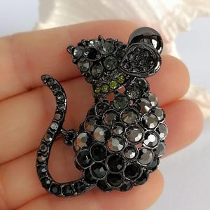 May include: A black and gray rhinestone mouse brooch. The brooch features a detailed design with a rounded body, long tail, and large ears. The mouse is adorned with various shades of gray and black rhinestones, with a few green rhinestones on the neck.