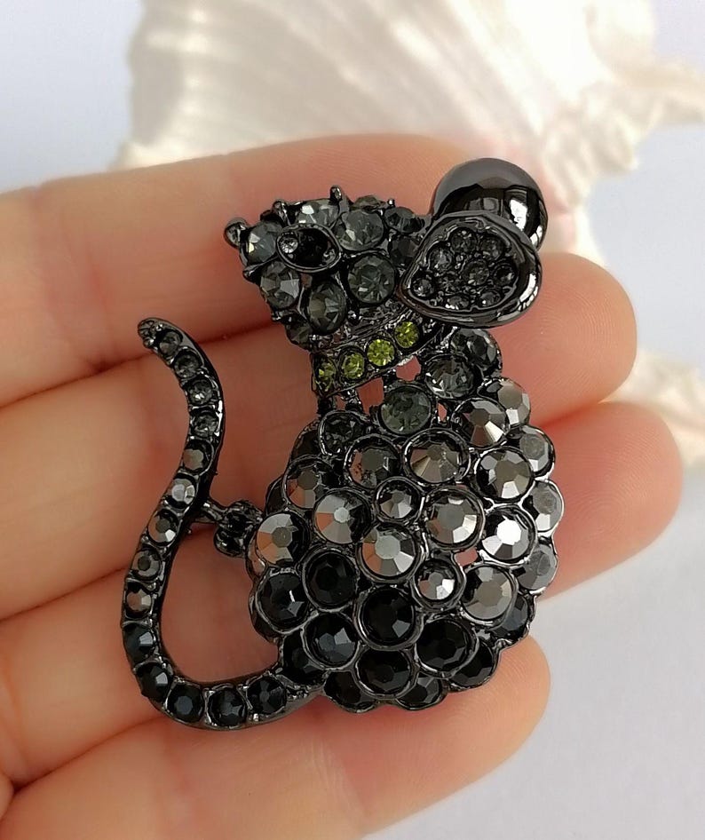 May include: A black and gray rhinestone mouse brooch. The brooch features a detailed design with a rounded body, long tail, and large ears. The mouse is adorned with various shades of gray and black rhinestones, with a few green rhinestones on the neck.