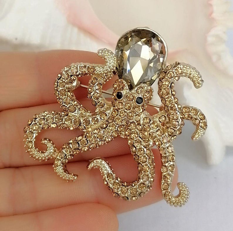 K&ouml;nnte beinhalten: Goldfarbene Oktopus-Brosche, besetzt mit kleinen, funkelnden Kristallen. Die Brosche hat einen gro&szlig;en, tropfenf&ouml;rmigen, facettierten Kristallkopf und schwarze Augen. Ein dekoratives Accessoire.
