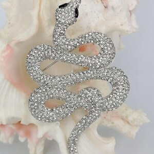 May include: A silver snake brooch, covered in clear crystals. The snake is in a coiled pose, with its head raised and black eyes. The brooch is displayed on a white and pink seashell.