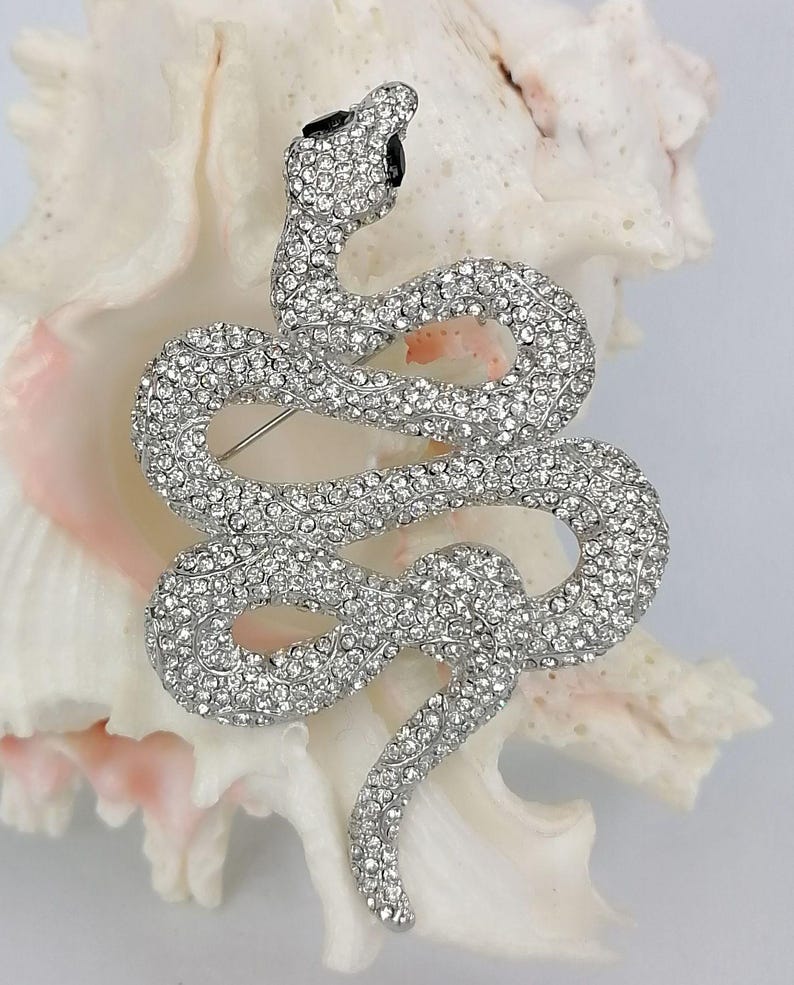 May include: A silver snake brooch, covered in clear crystals. The snake is in a coiled pose, with its head raised and black eyes. The brooch is displayed on a white and pink seashell.