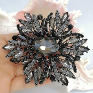 Op de afbeelding: Een grote, ronde broche met een centrale ovaalvormige donkergrijze kristal, omringd door een stervormige schikking van marquise-geslepen grijze kristallen, kleine ronde zwarte kralen en heldere strass-steentjes in een donkerkleurig metalen montuur.
